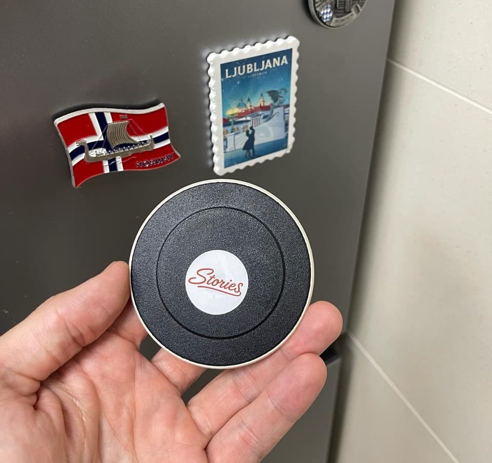 A hand holding a travel magnet with a Stories sticker on it, in front of a fridge